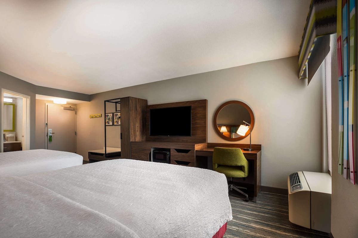 A hotel room with two beds, a desk, a flat-screen TV, a mirror, and a small heater.