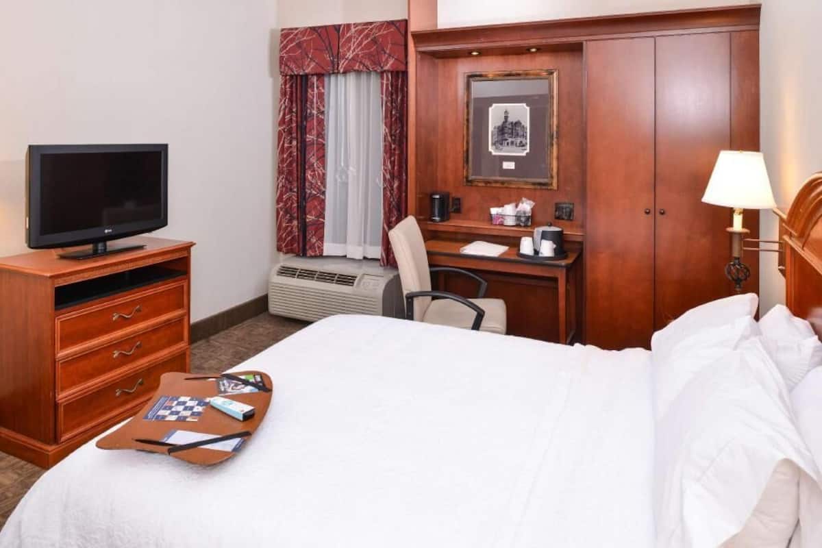 A hotel room with a bed, a desk with a chair, a television, a desk lamp, and a framed picture on the wall.