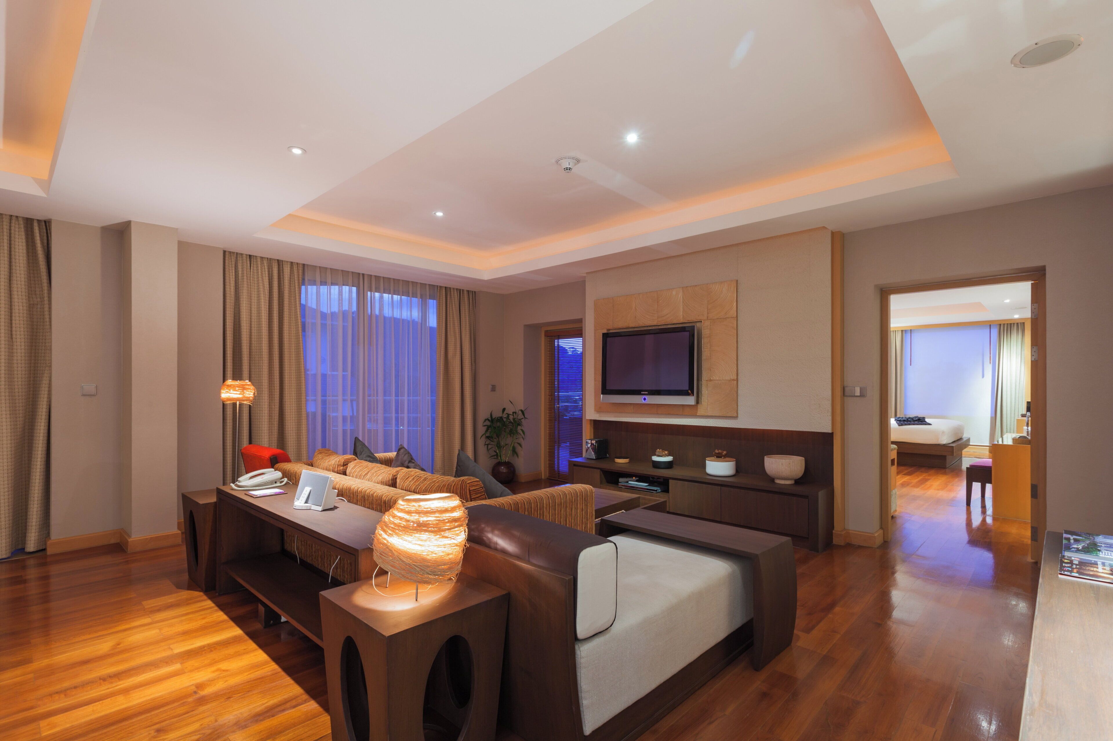 A modern hotel room with a wooden floor, a sofa set, a dining table, a TV mounted on the wall, and a view of the city through the window.