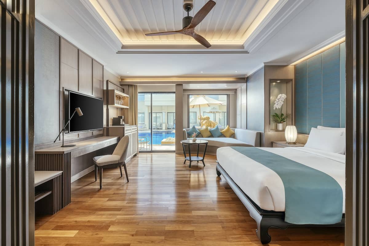 A modern hotel room with a large bed, a desk with a TV, a seating area with a table and chairs, and a view of a pool through large windows.