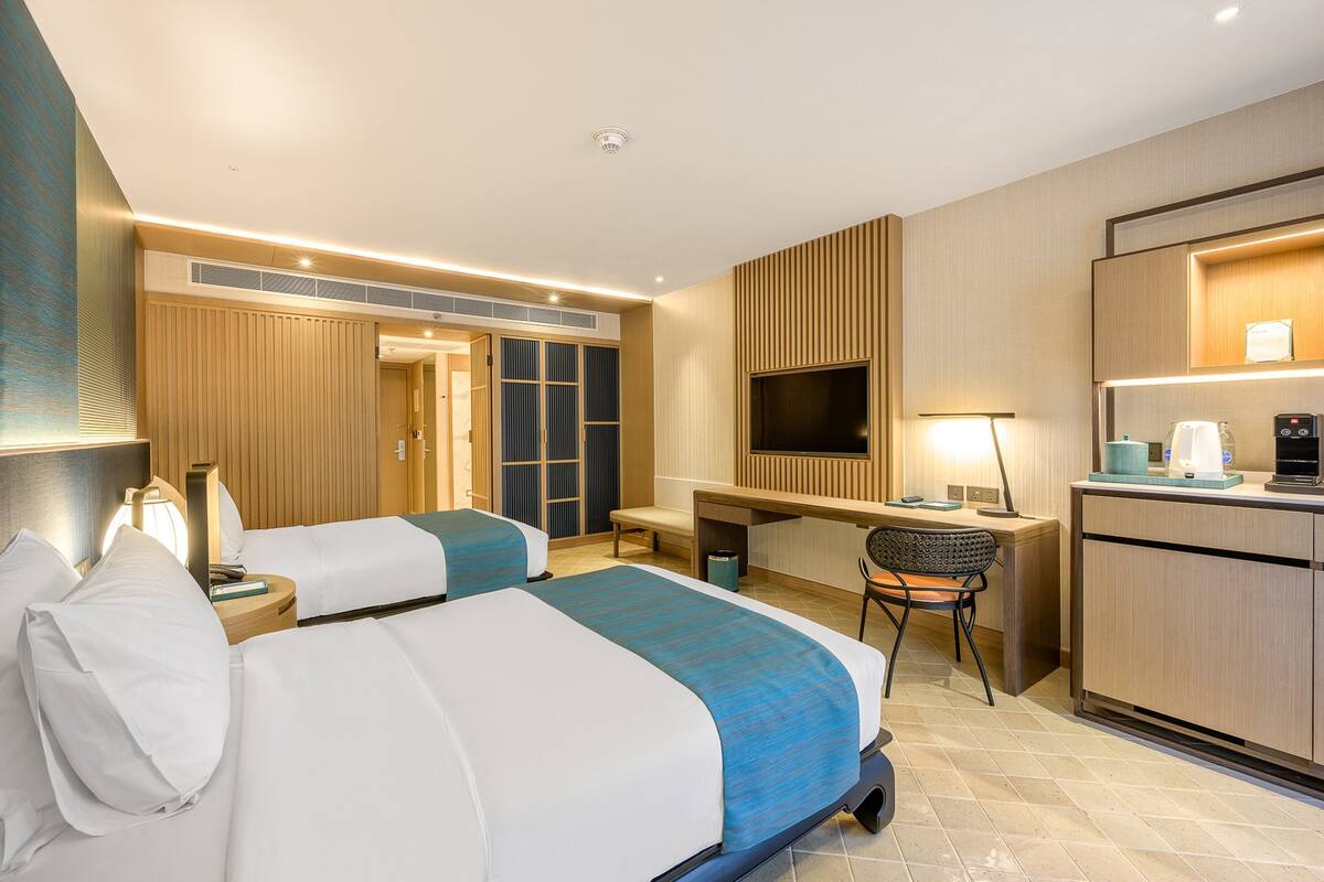 A hotel room with two beds, a desk, a chair, a TV, and a kitchenette.
