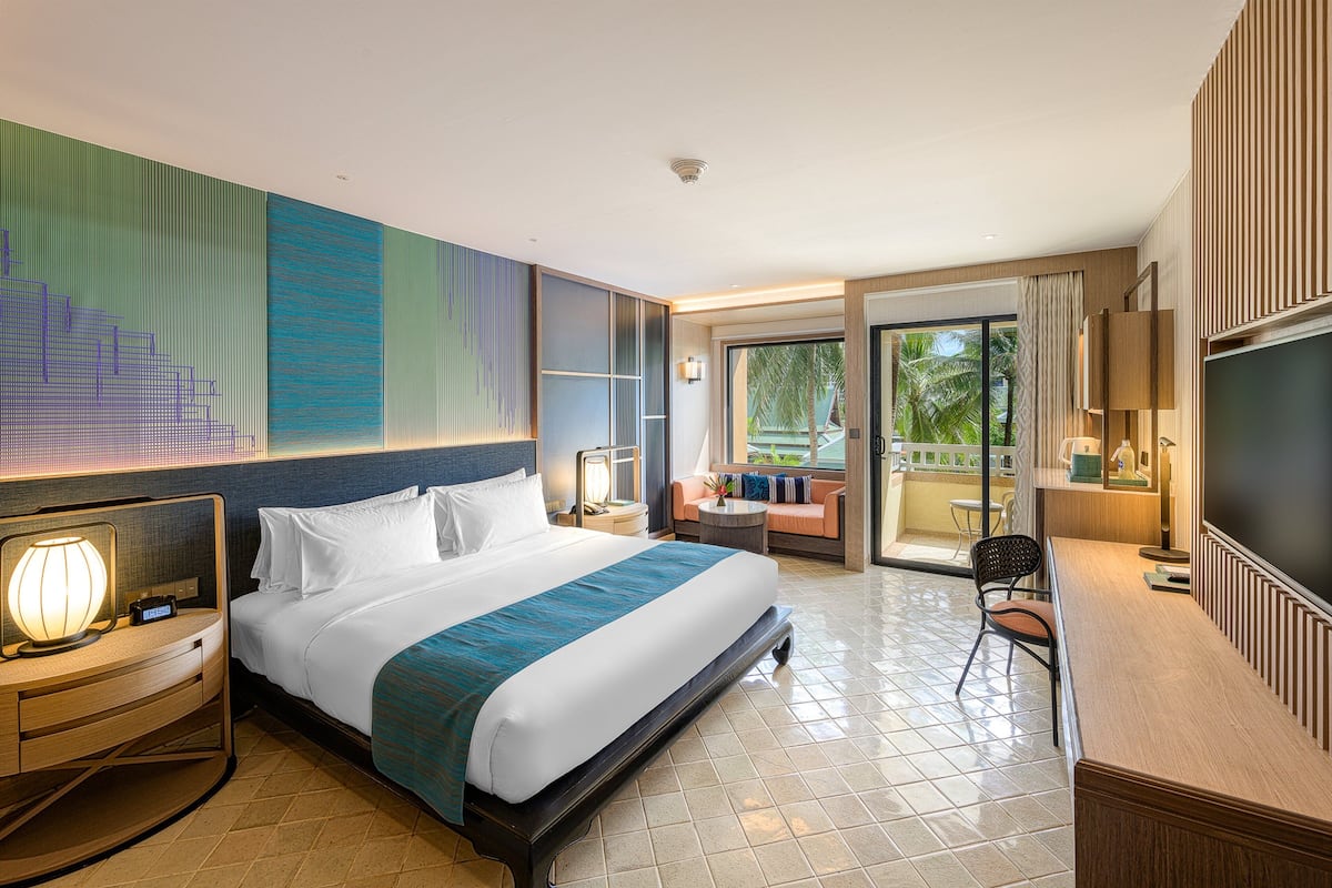 A hotel room with a large bed, a desk with a chair, a TV, and a balcony with a view of palm trees.