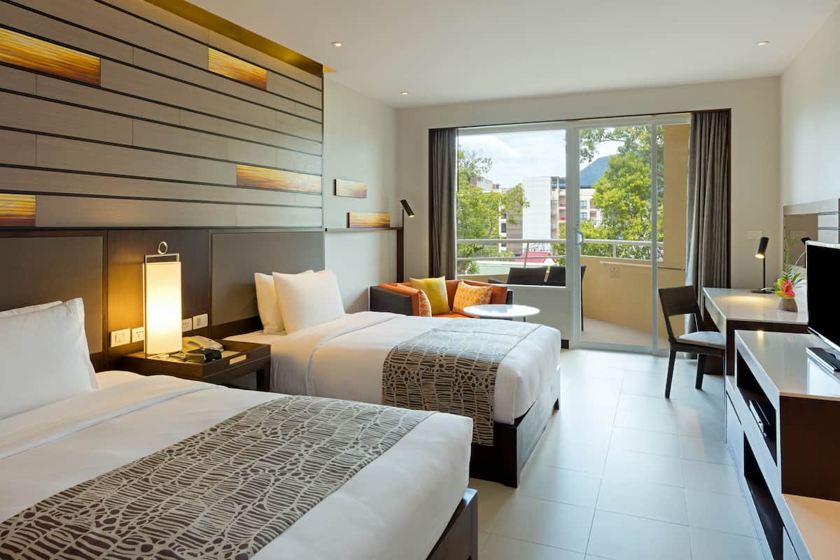 A hotel room with two beds, a desk, a chair, and a TV. There is a balcony with a view of buildings and trees.