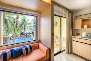 A hotel room with a view of a swimming pool, a seating area with cushions, a kitchenette, and a sliding glass door to the balcony.