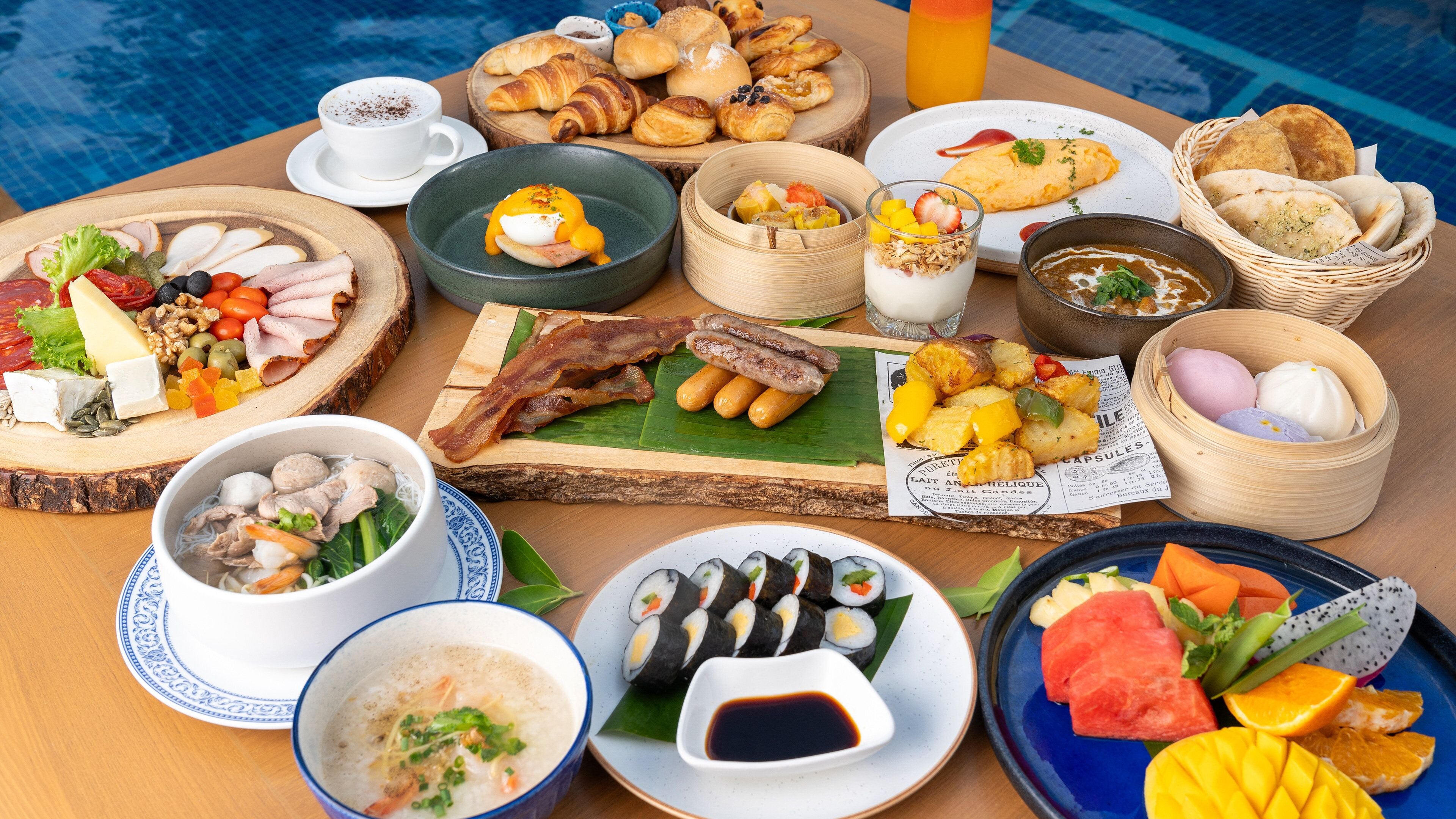 A variety of breakfast foods including pastries, meats, and fruits, arranged on a wooden table near a pool.