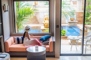 A room with a sofa, a round table, and a view of an outdoor swimming pool and garden.