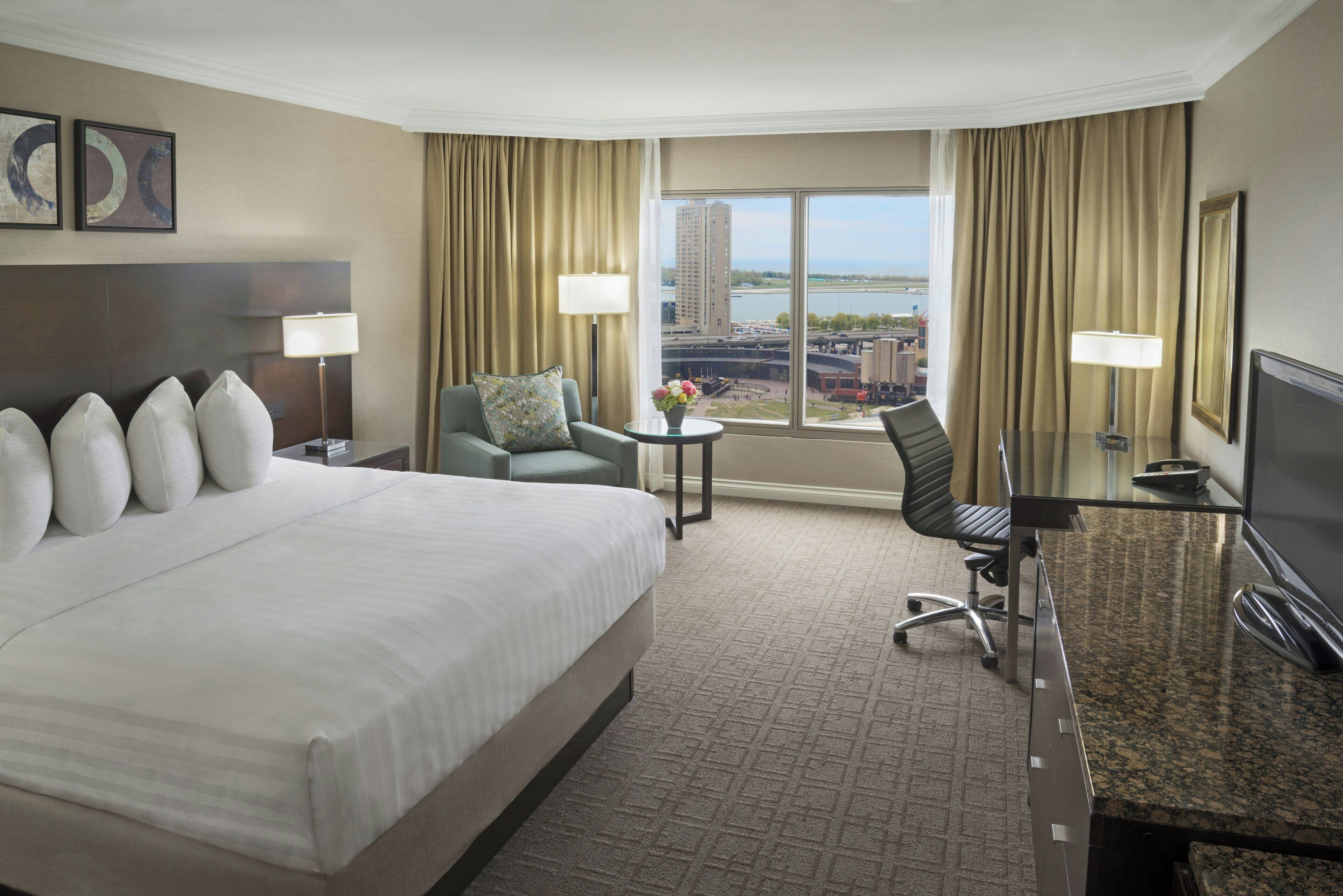A hotel room with a large bed, a desk, a chair, a small table with a vase, and a view of a cityscape through the window.