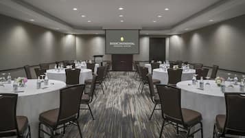 Un salón de conferencias con varias mesas, sillas y una pantalla para presentaciones al frente.