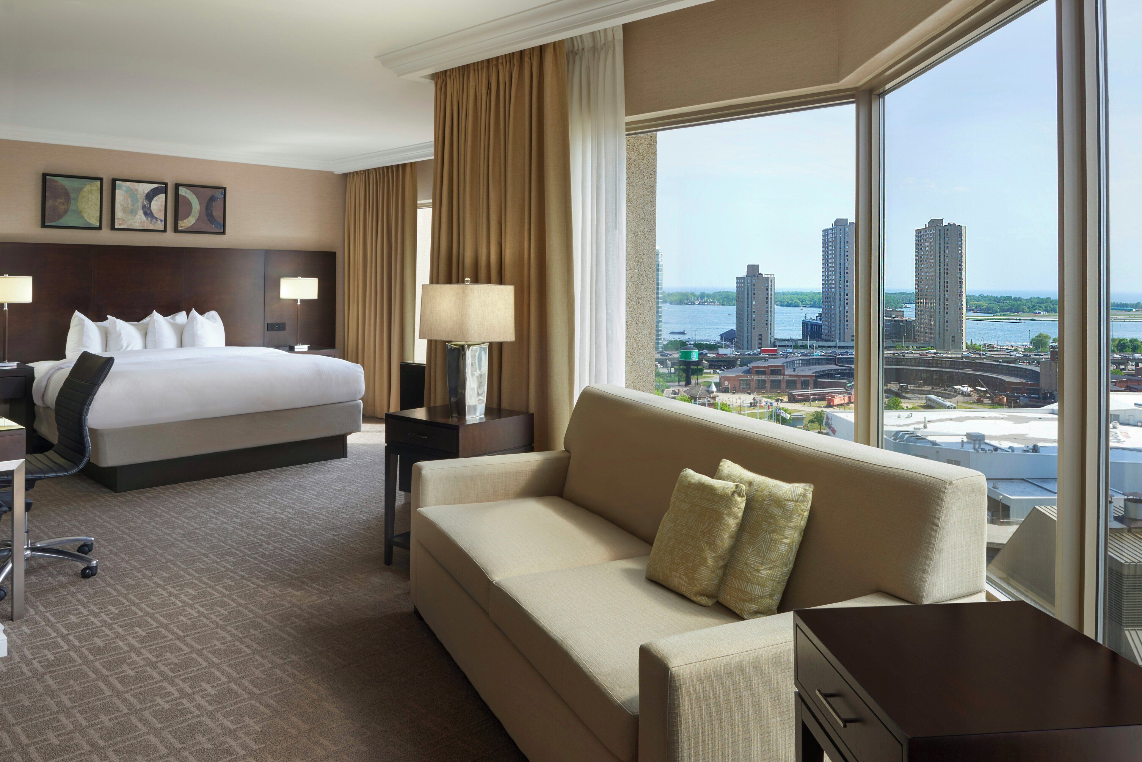 A hotel room with a bed, a sofa, a desk, and a large window offering a city view.