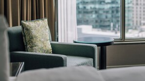 Habitación de hotel con vista a un paisaje urbano a través de ventanas amplias, un sofá con un cojín estampado y una lámpara de pie.