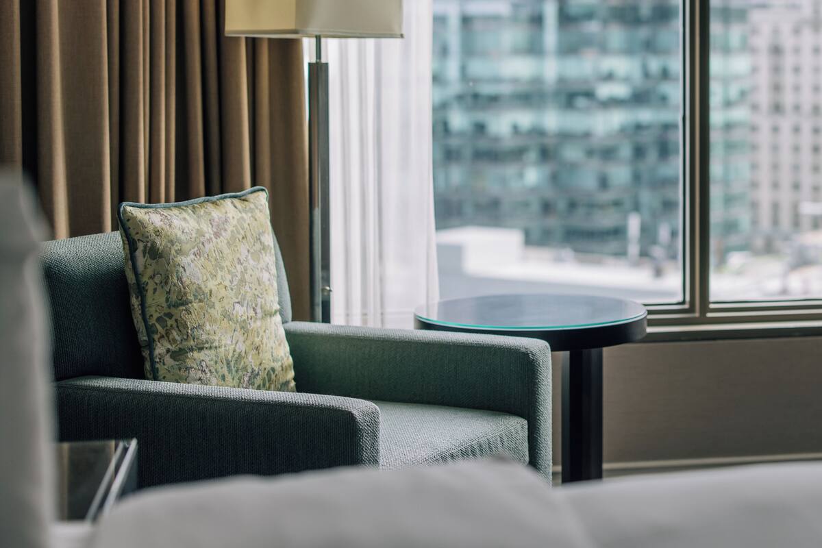 A hotel room with a view of a cityscape through large windows, a sofa with a patterned pillow, and a floor lamp.