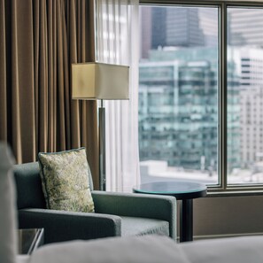 A hotel room with a view of a cityscape through large windows, a sofa with a patterned pillow, and a floor lamp.
