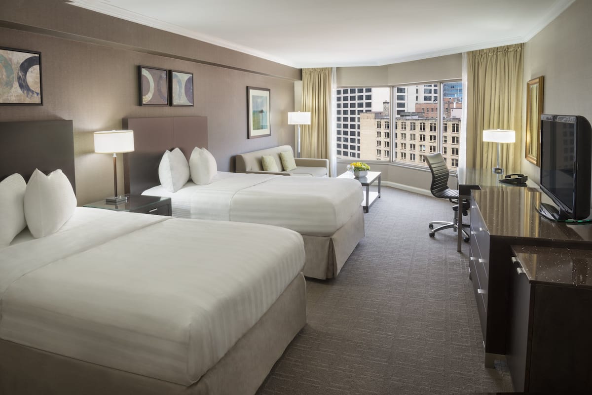 A hotel room with two beds, a desk, a TV, and a window with a city view.