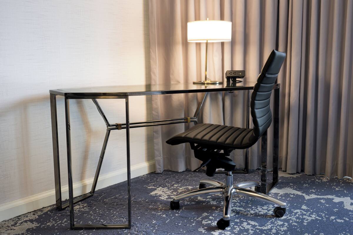 A modern office chair with a high backrest and armrests, positioned next to a glass desk with metal legs. A desk lamp is placed on the desk, and the room features a carpeted floor.