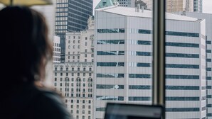 Una persona trabajando en un escritorio con una computadora portátil, con vista a un paisaje urbano de edificios modernos y una torre distintiva con el logo 'TD'.