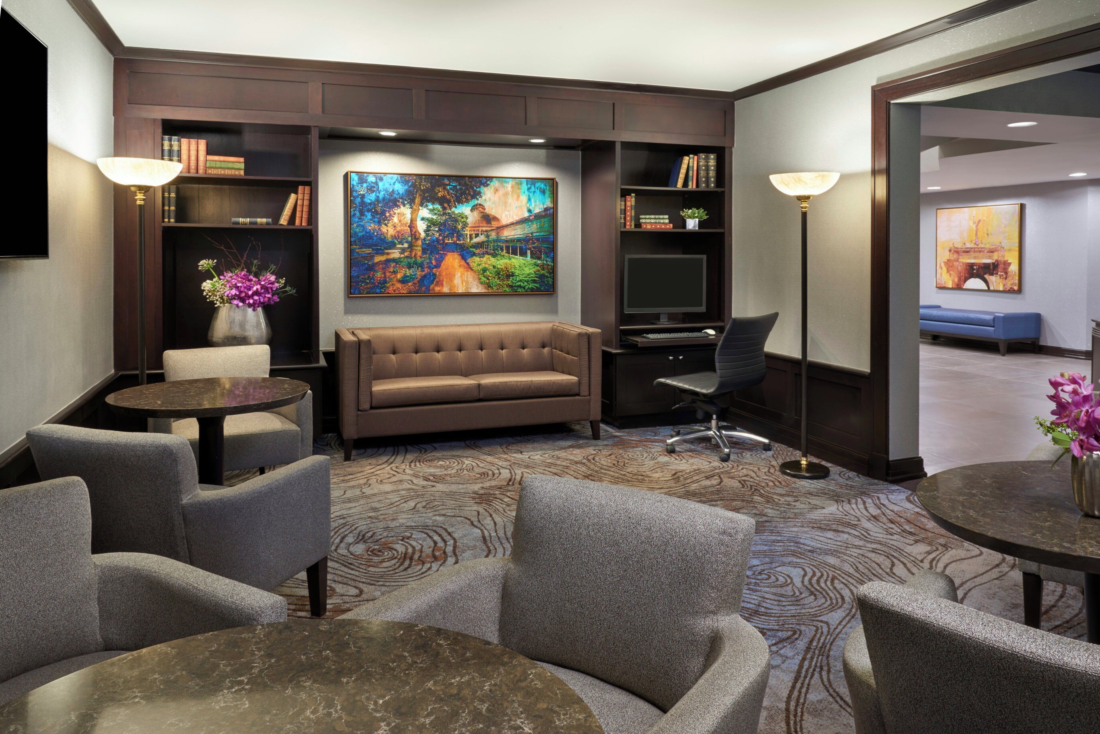 Quarto de hotel com sofá, poltronas, escrivaninha, televisão e uma pintura na parede.