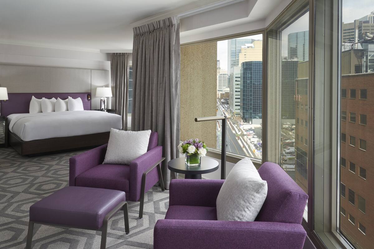 A hotel room with a bed, two purple armchairs, a small table with a vase of flowers, and a large window with a city view.