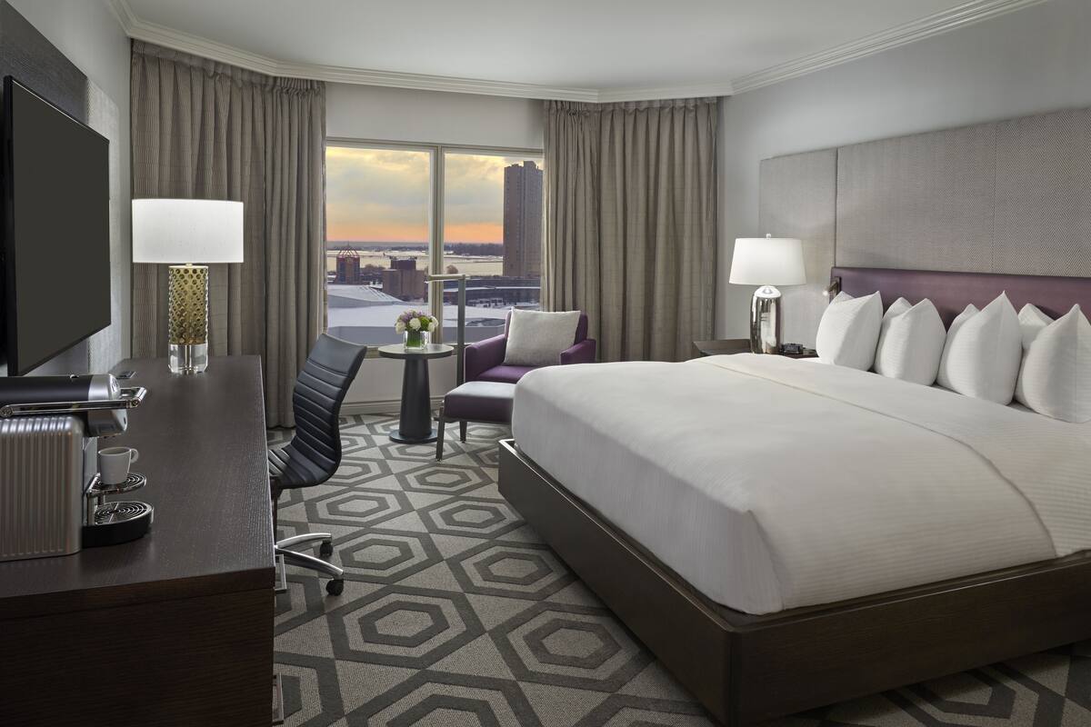 A hotel room with a large bed, a desk with a coffee maker, a TV, and a view of the cityscape.