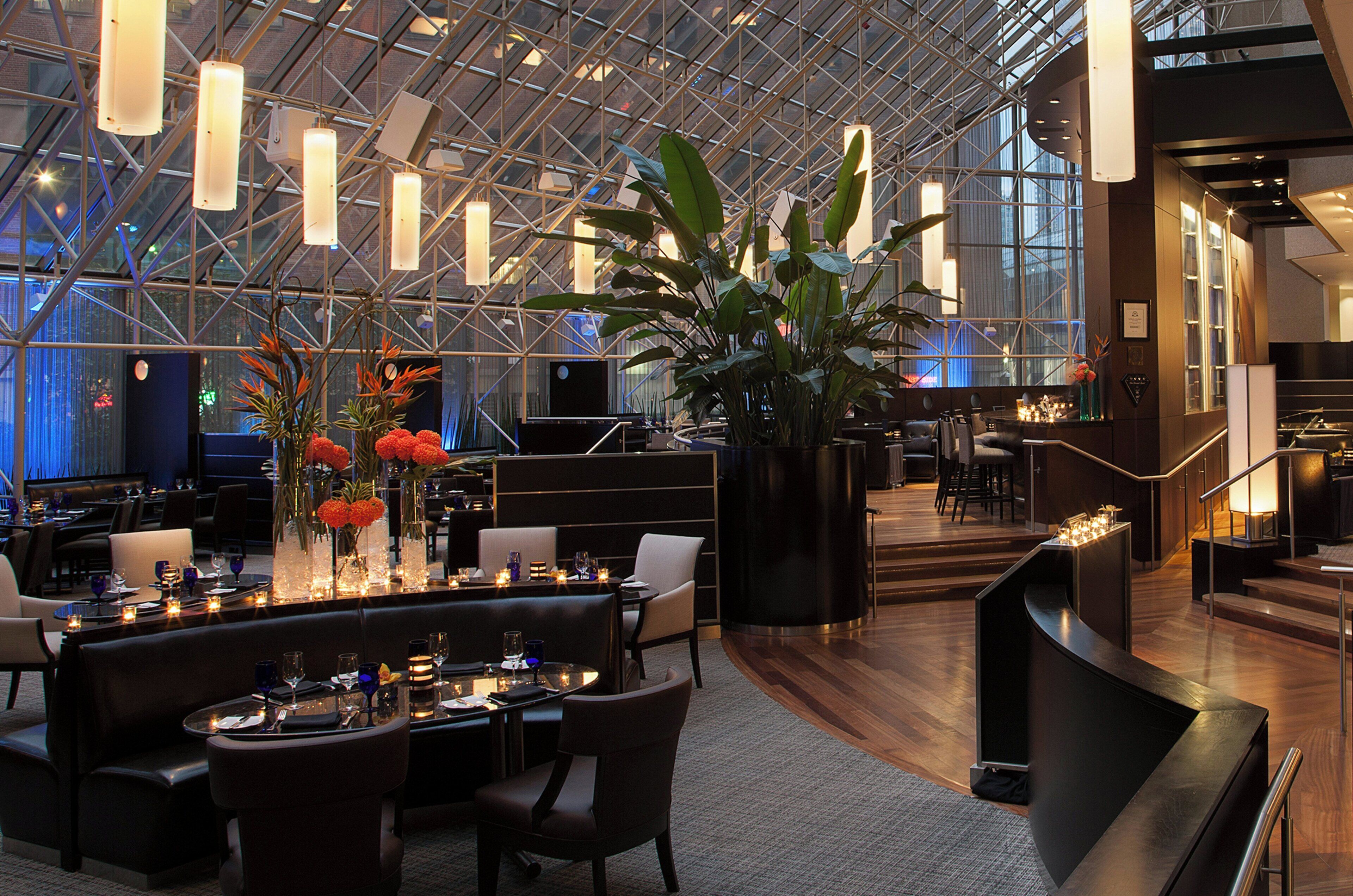 Um lobby moderno de hotel com teto alto, paredes de vidro e área de estar central com mesas e cadeiras.