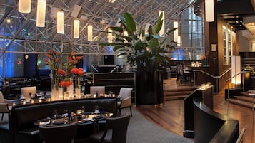 Um lobby moderno de hotel com teto alto, paredes de vidro e área de estar central com mesas e cadeiras.