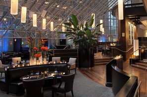 Un moderno hall de hotel con techo alto, paredes de vidrio y una zona central de estar con mesas y sillas.