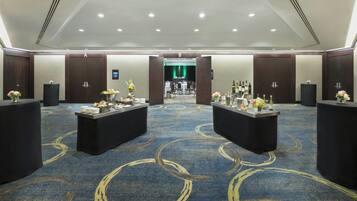 A spacious hotel lobby with a blue carpet featuring circular patterns, two reception desks with floral centerpieces, and a green-lit stage in the background.
