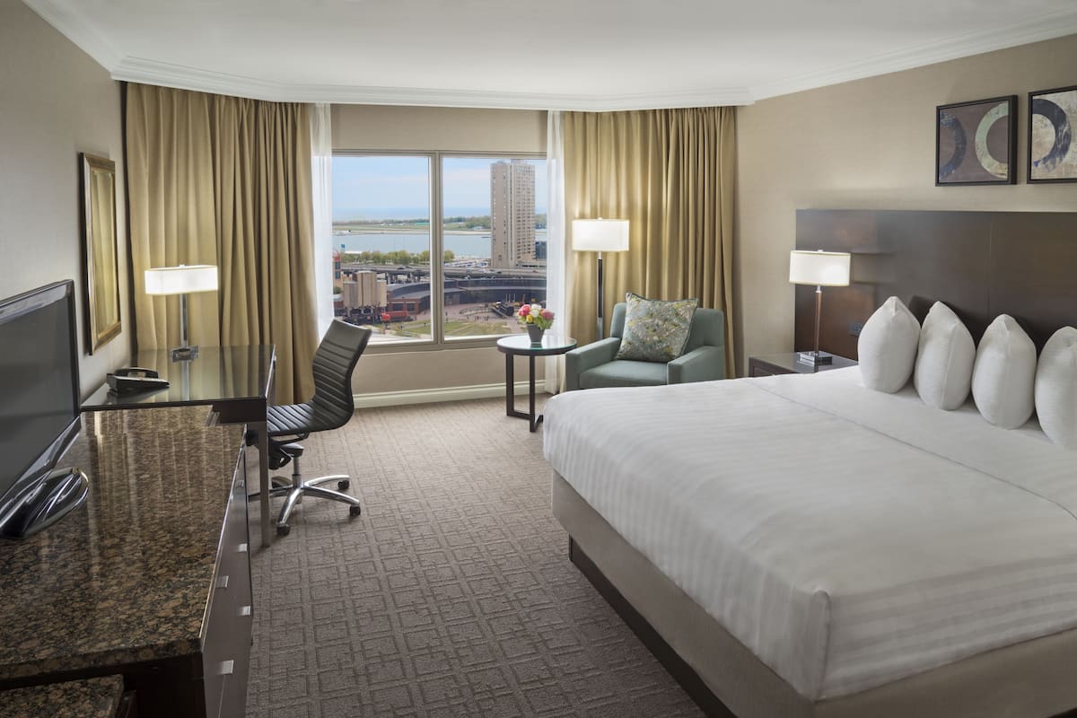 A hotel room with a large bed, a desk with a computer, a chair, a small table with a lamp, and a view of a cityscape through large windows.