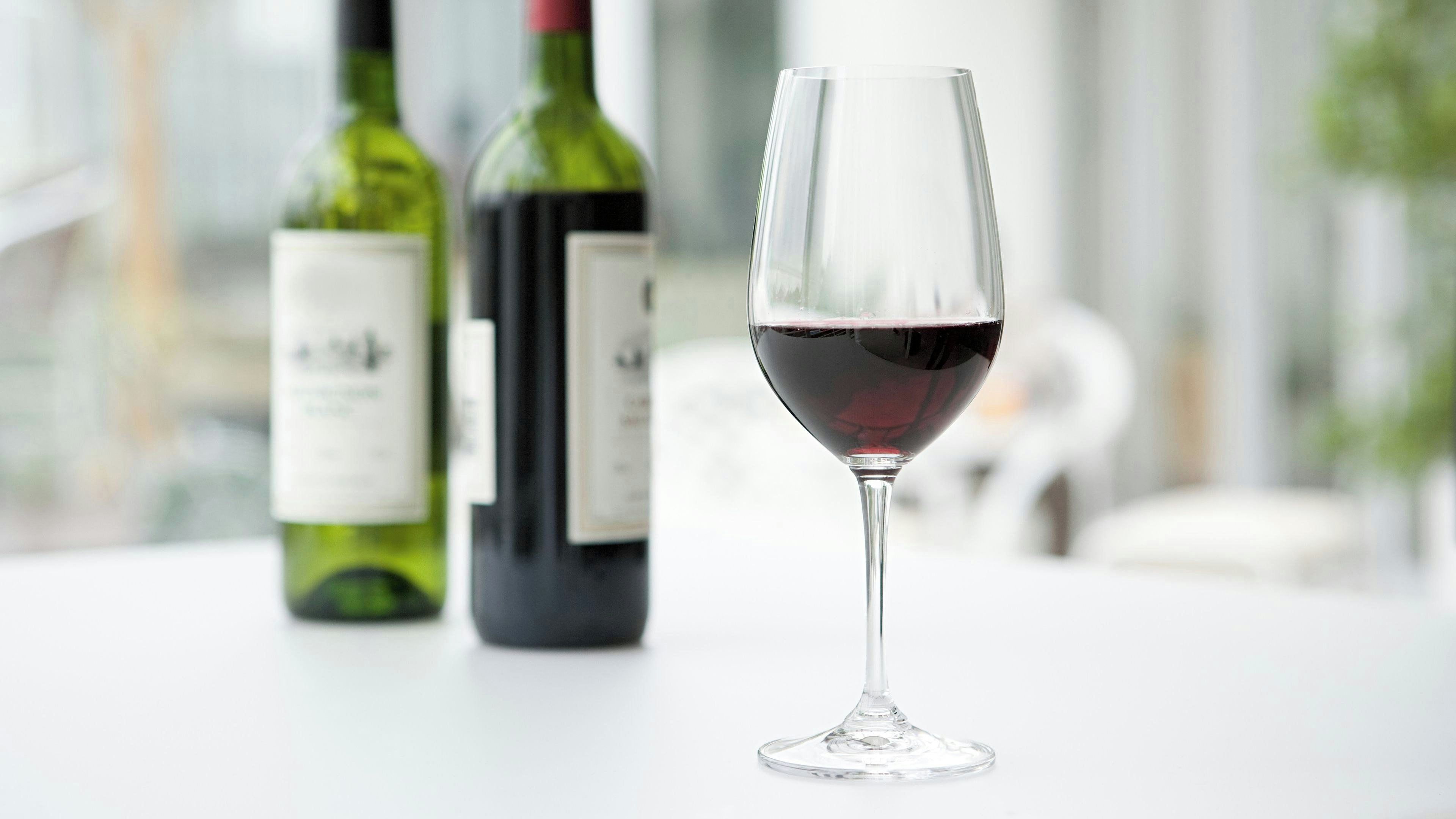 A wine glass with red wine on a table, with two wine bottles in the background.