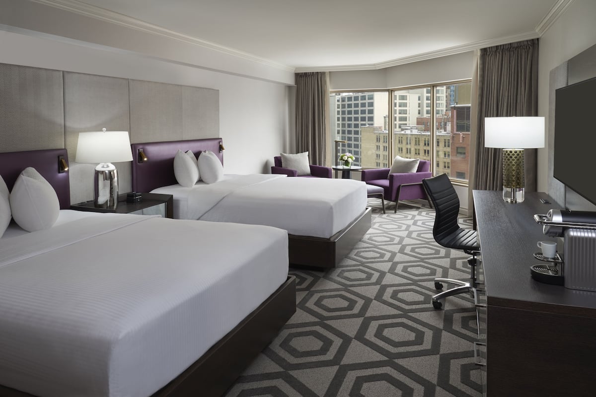 A hotel room with two beds, a desk with a coffee maker, a TV, and a view of the city.