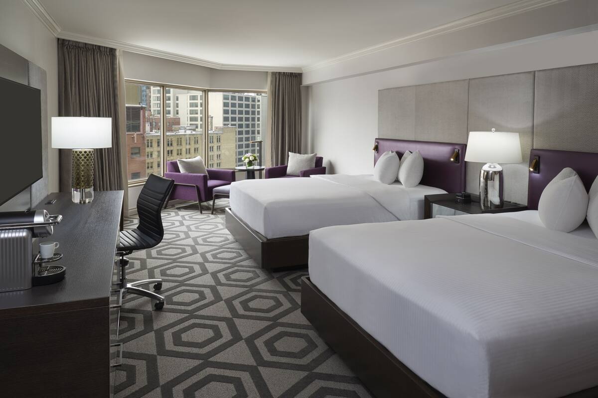 A hotel room with two beds, a desk, a chair, and a view of the cityscape.