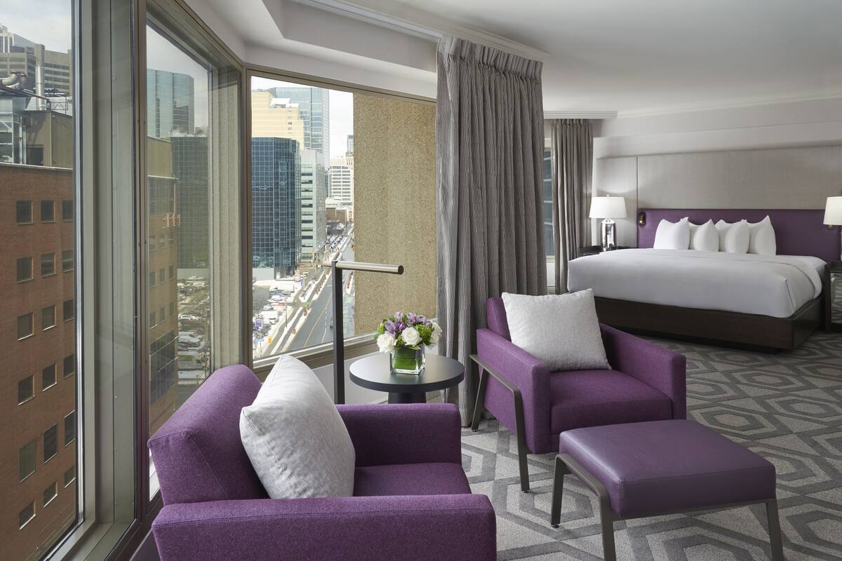 A hotel room with a large bed, two purple armchairs, a small table with a vase of flowers, and a view of the city through large windows.
