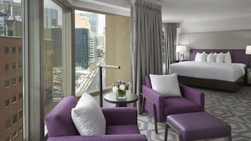 Una habitación de hotel con una cama grande, dos sillones morados, una mesita con un jarrón de flores y vistas a la ciudad a través de amplios ventanales.