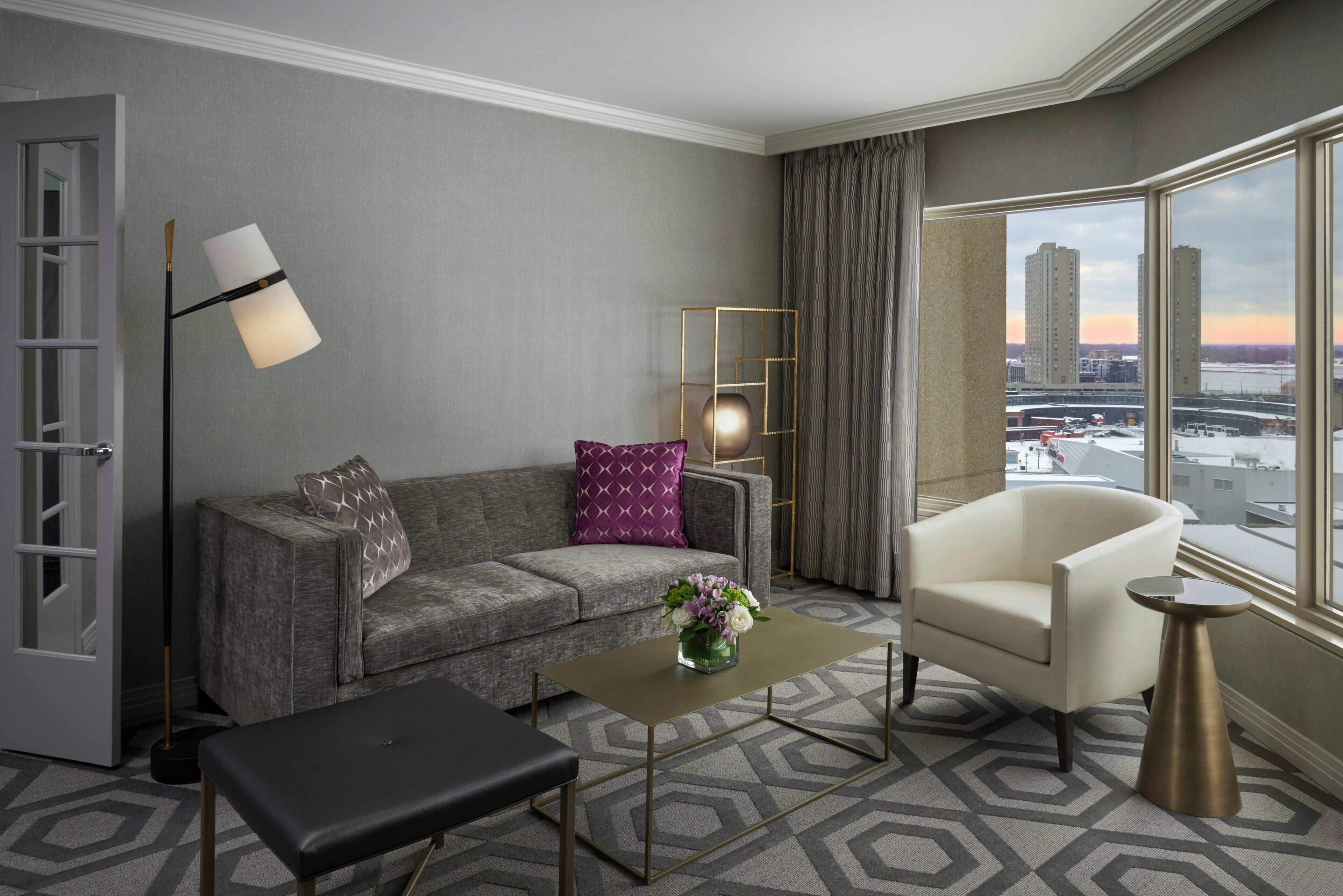 Une chambre d’hôtel moderne avec un canapé gris, un fauteuil blanc, une petite table et une vue sur le paysage urbain à travers de grandes fenêtres.