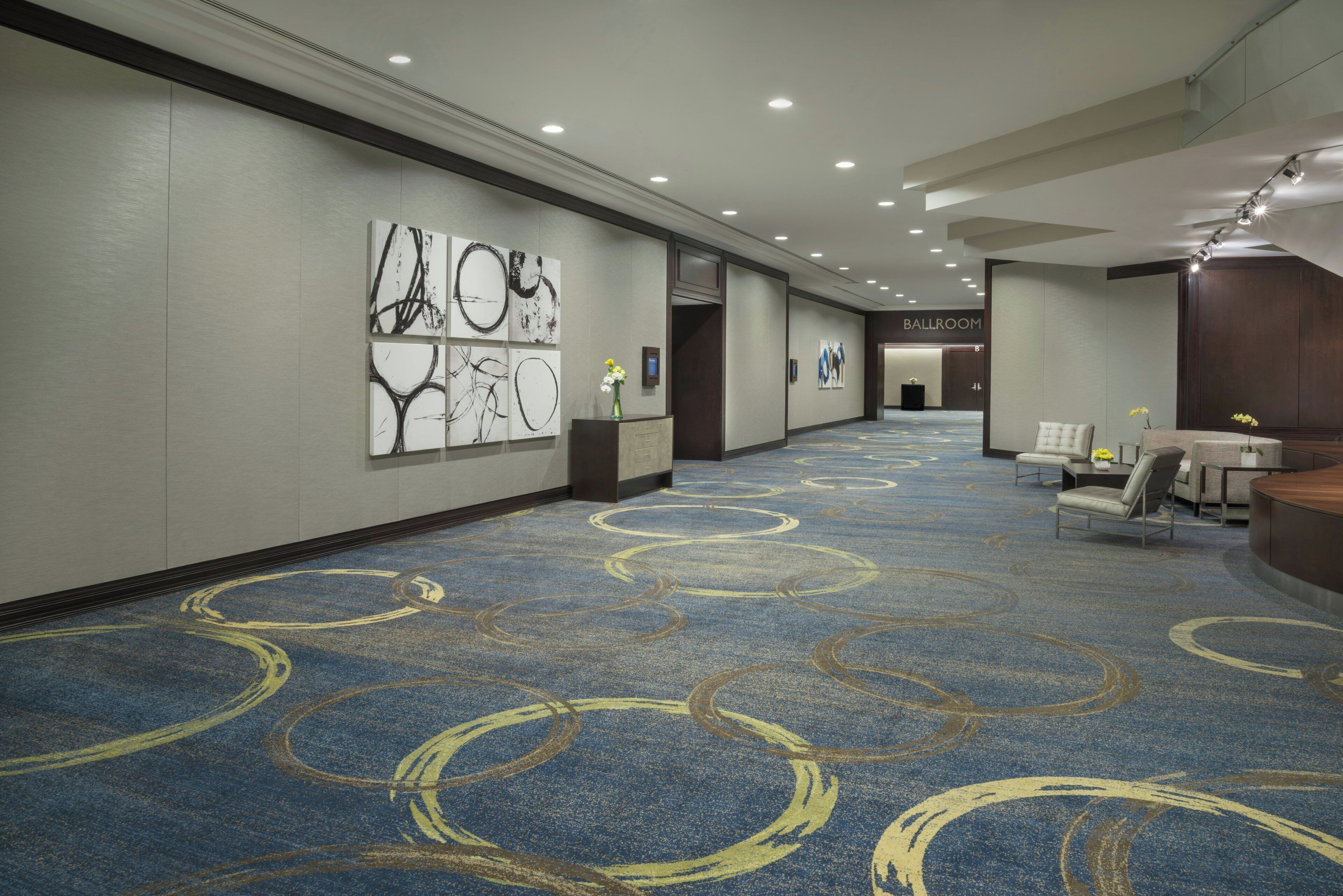 Een hotelgang met een blauw tapijt met cirkelvormige patronen, een receptiegedeelte met een balie en zitplaatsen, en kunstwerken aan de muren.