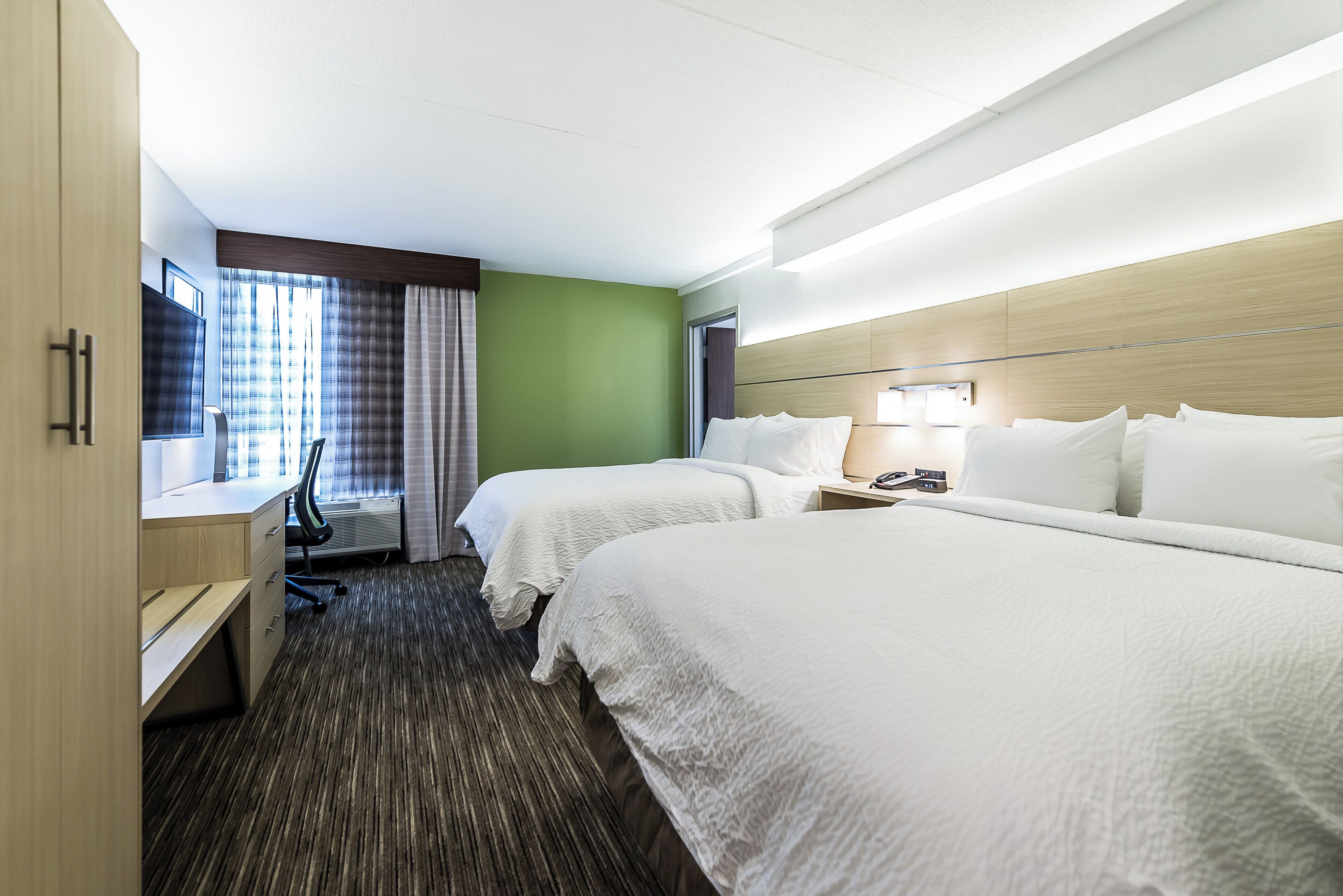A hotel room with two beds, a desk, a chair, a window with curtains, and a bedside table with a lamp.