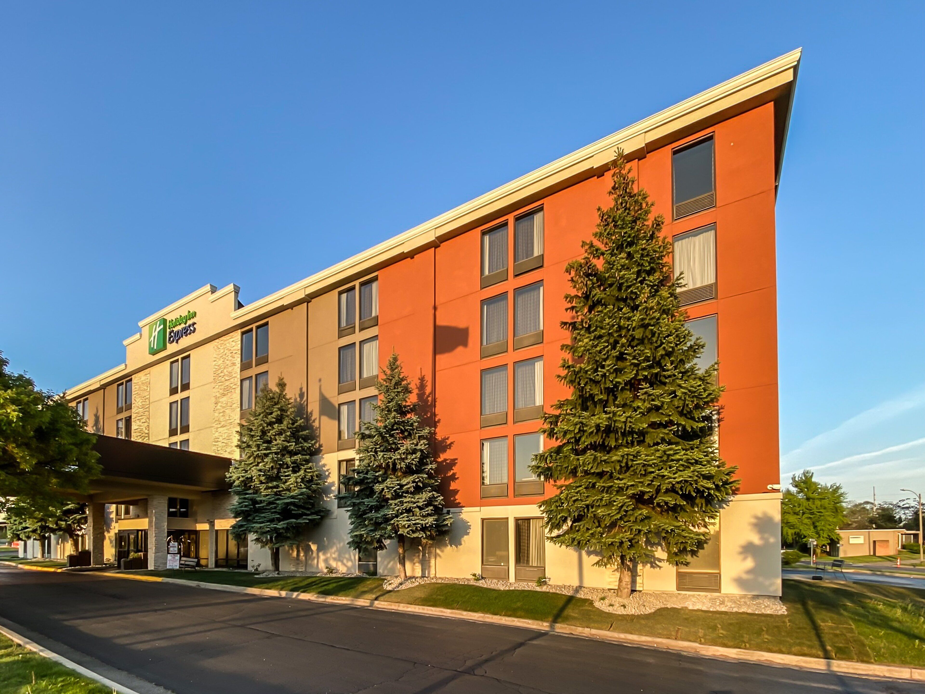 A modern hotel with a red and beige facade, multiple windows, and three evergreen trees in front.
