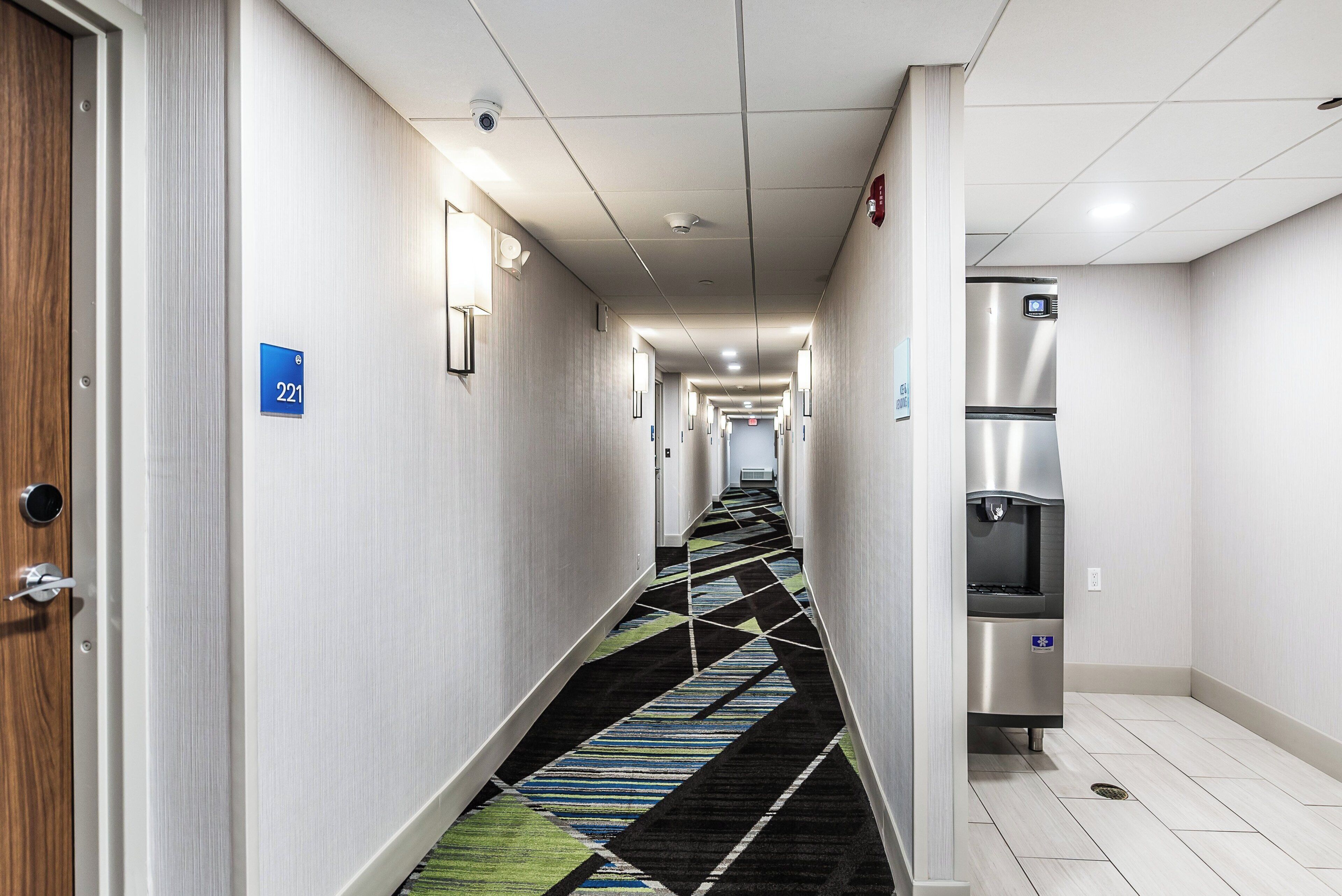 A hotel hallway with a carpeted floor, a refrigerator, and a fire alarm on the wall.