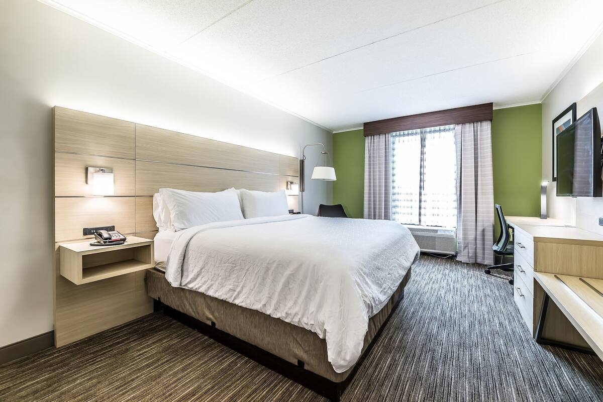 Une chambre d’hôtel comprenant un lit, une table de chevet avec un téléphone, un bureau avec une chaise, une télévision et une fenêtre avec des rideaux.