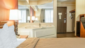 A hotel room with a large bed, a sitting area with a chair and table, a large mirror, a bathroom with a bathtub, and a door leading to another room. - Quality Inn Gaylord (Gaylord)