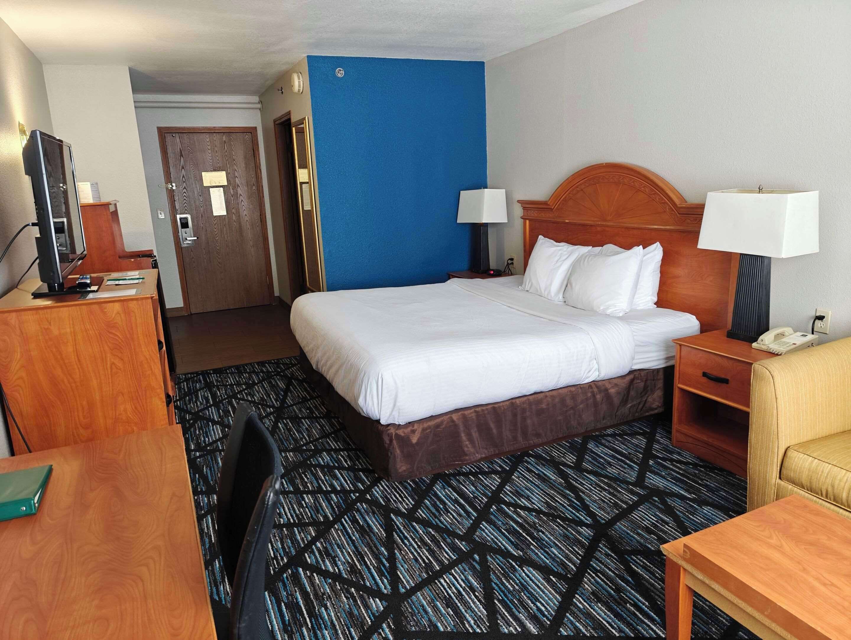 A hotel room with a large bed, two bedside lamps, a desk with a computer, a chair, and a small table.