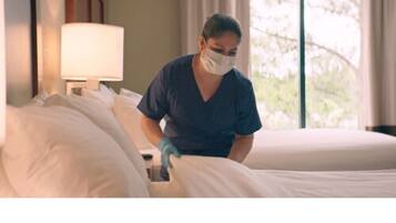 A person in a dark blue uniform cleaning a bed in a room with a window and a lamp on a bedside table.