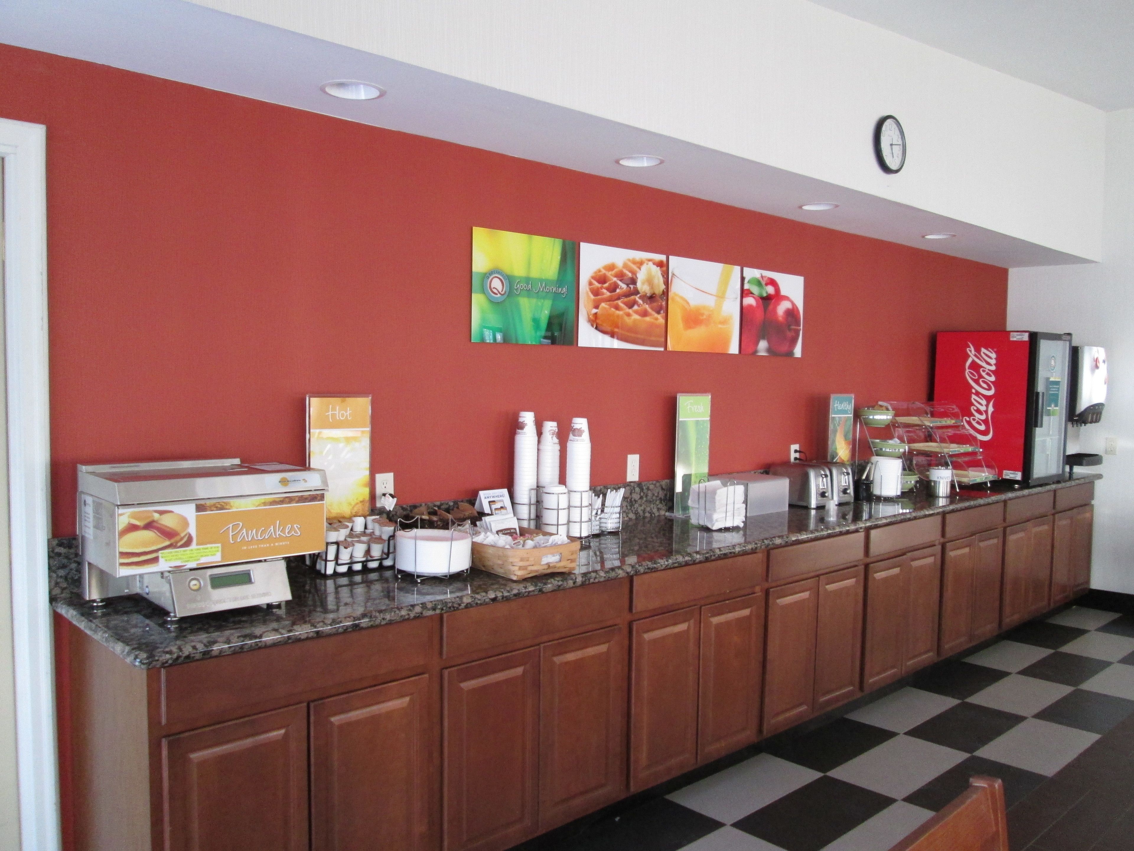 Una barra de desayuno con diversas opciones de comida y bebida, que incluye una estación de alimentos calientes, zumos recién exprimidos y un refrigerador de Coca-Cola.