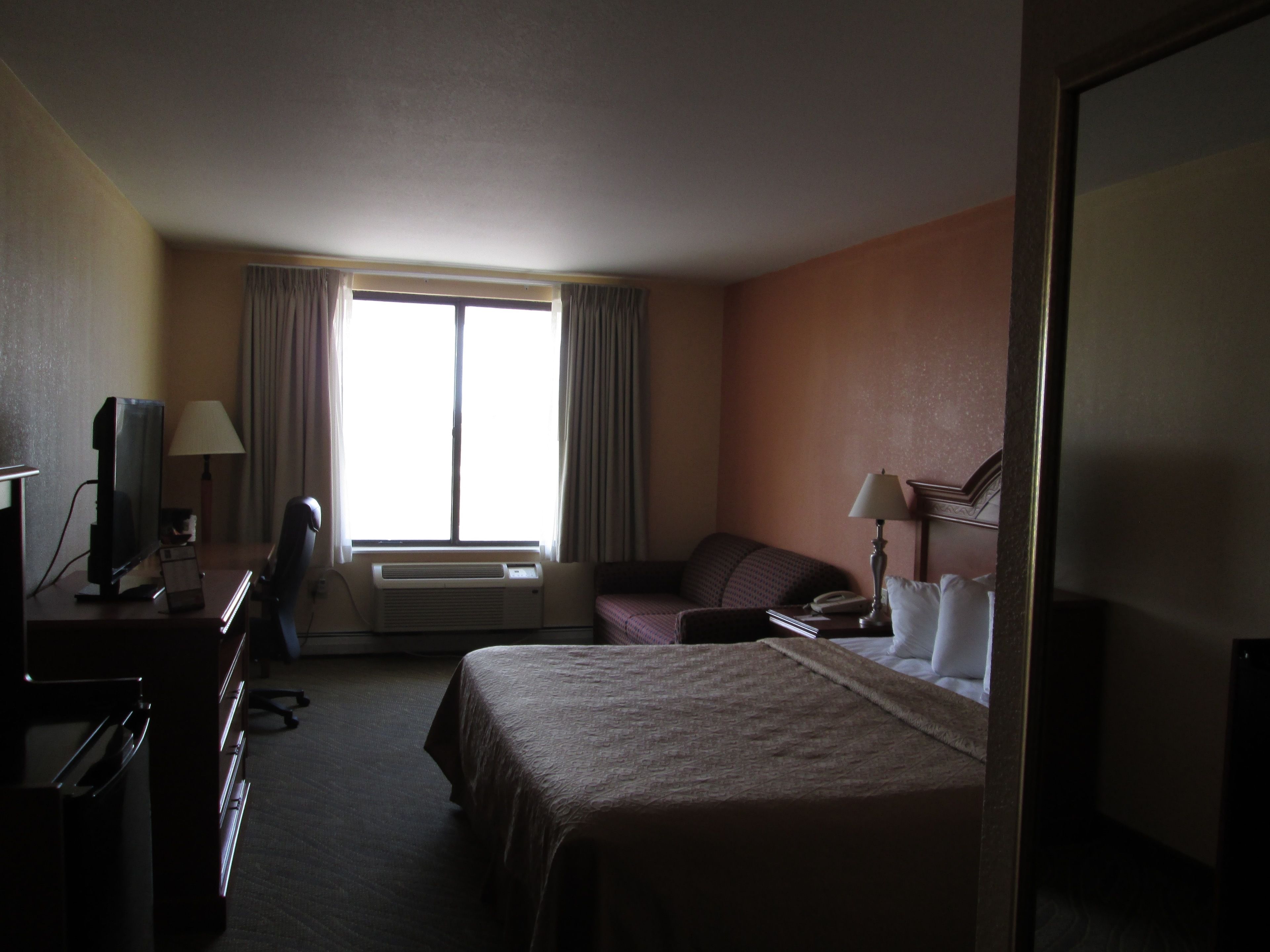 A hotel room with a bed, a desk with a computer, a chair, a window with curtains, and a wall-mounted air conditioner.