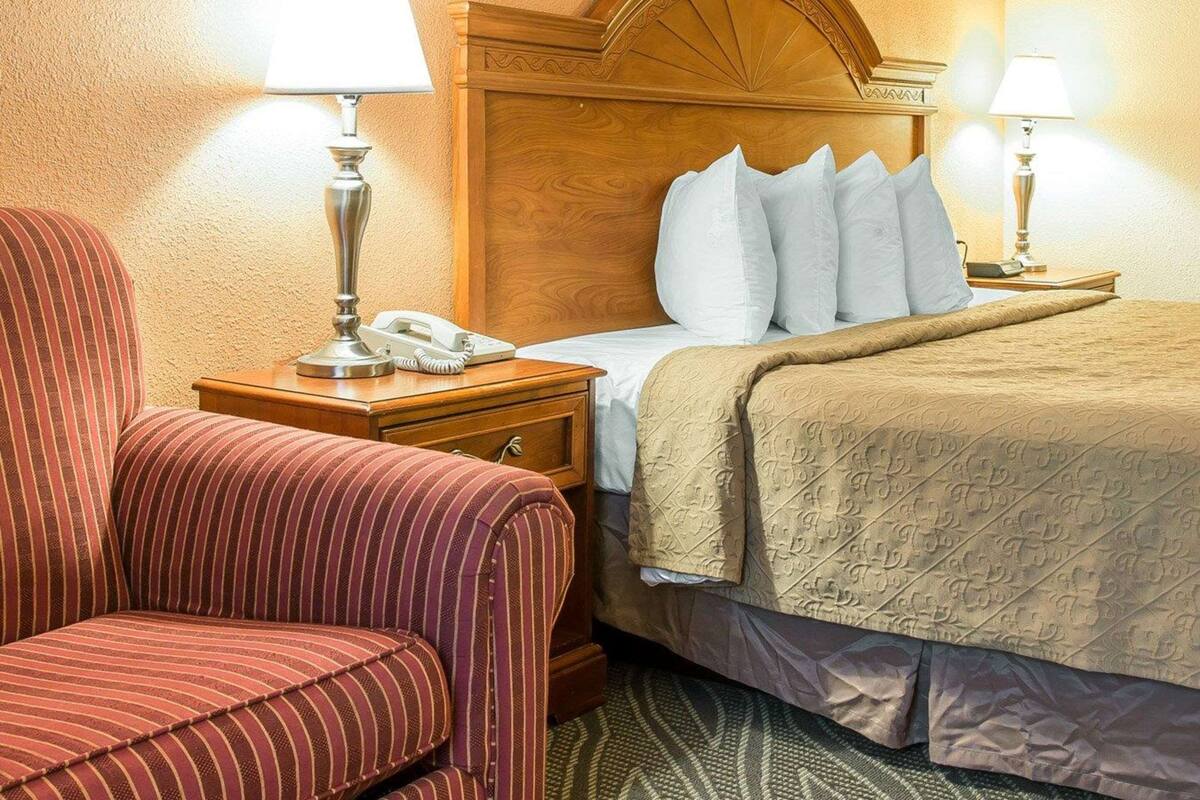 Camera d'albergo con testiera in legno, una poltrona a righe, un comodino con lampada, un telefono e un letto con coperta beige.