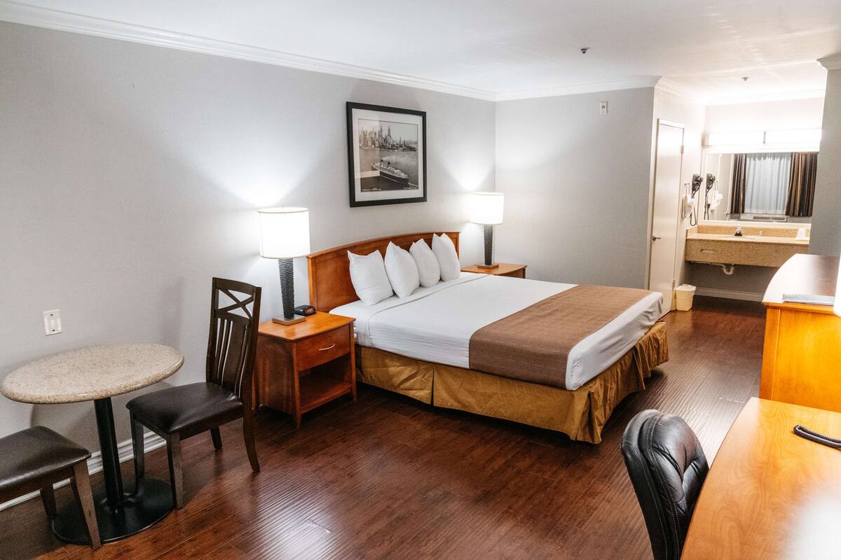 Hotellihuone, jossa on sänky, kaksi yöpöytää, tuoli, pieni pyöreä pöytä, peili ja taulu seinällä.