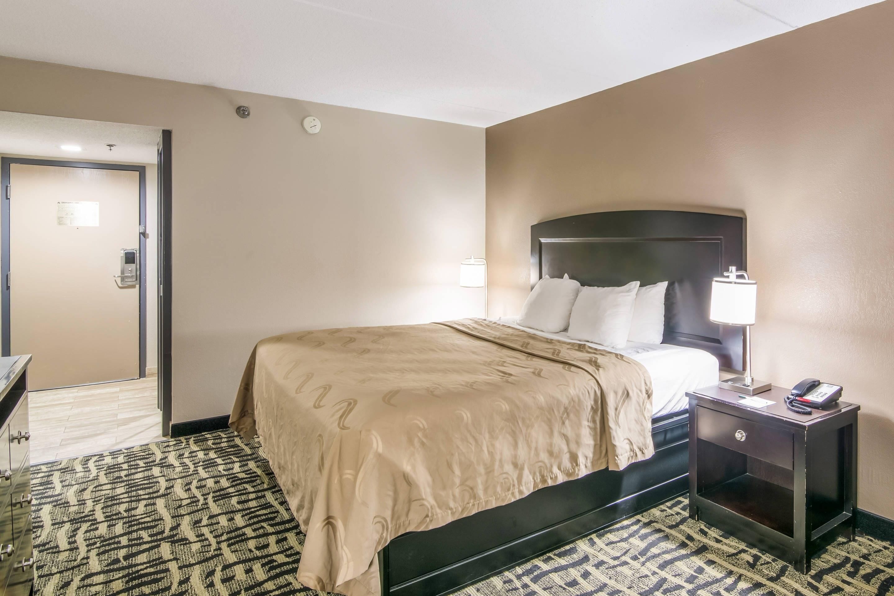 A hotel room with a bed, bedside table, lamp, and a door leading to another room.