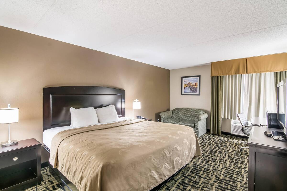 Ein Hotelzimmer mit einem großen Bett, einem Schreibtisch mit Computer, einem Sessel, einer Lampe und einem Bild an der Wand.