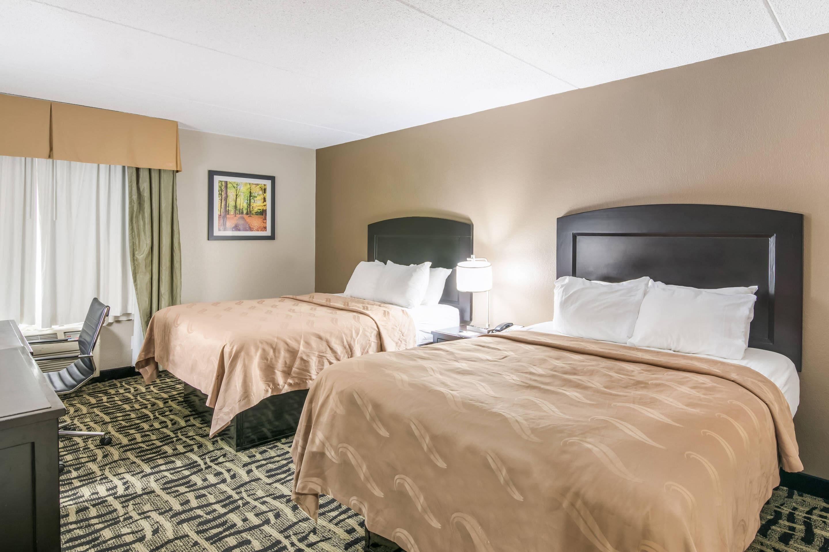 A hotel room with two beds, a desk, a chair, a lamp, and a framed picture on the wall.