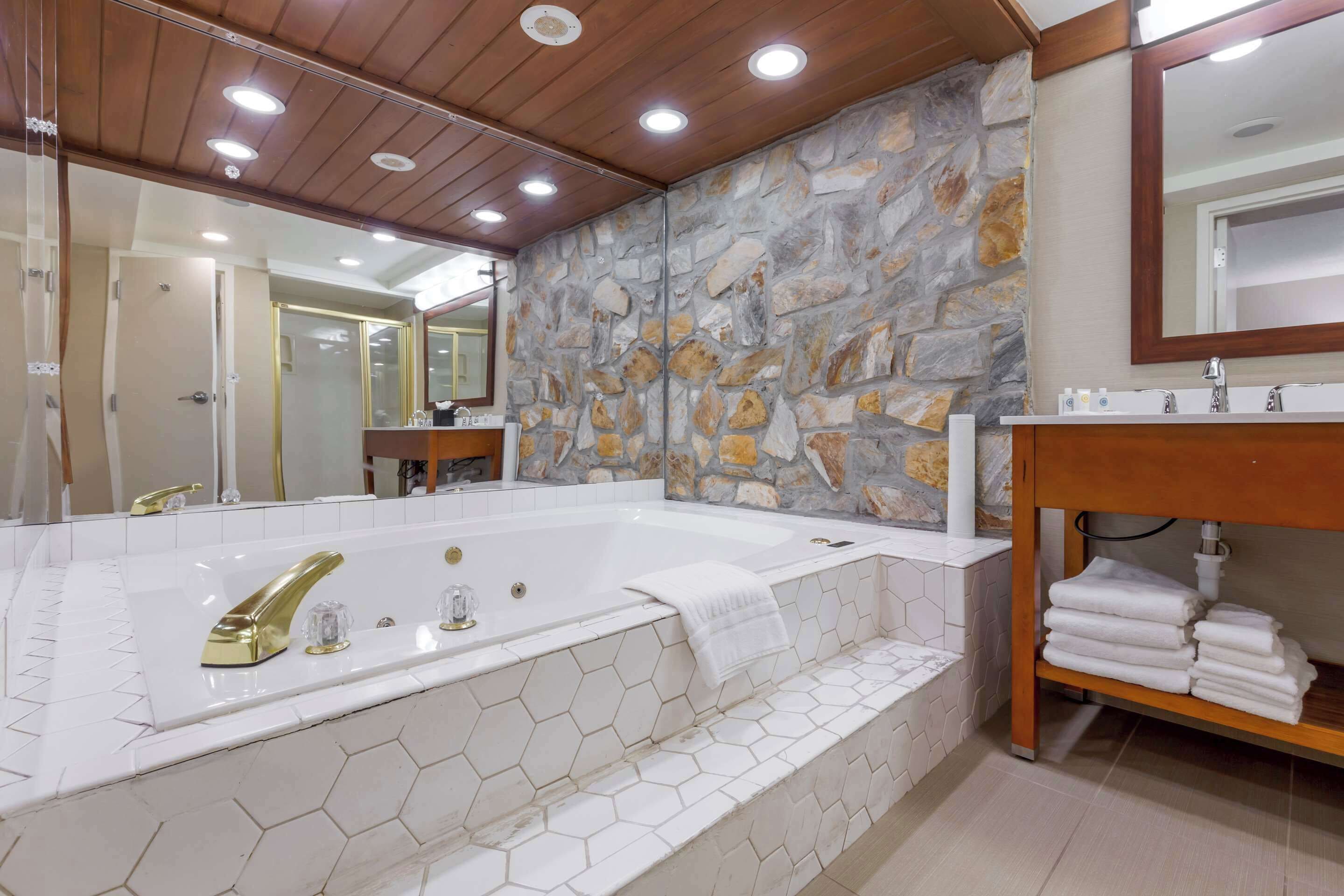 A bathroom with a large bathtub, a wooden vanity, and a stone wall.