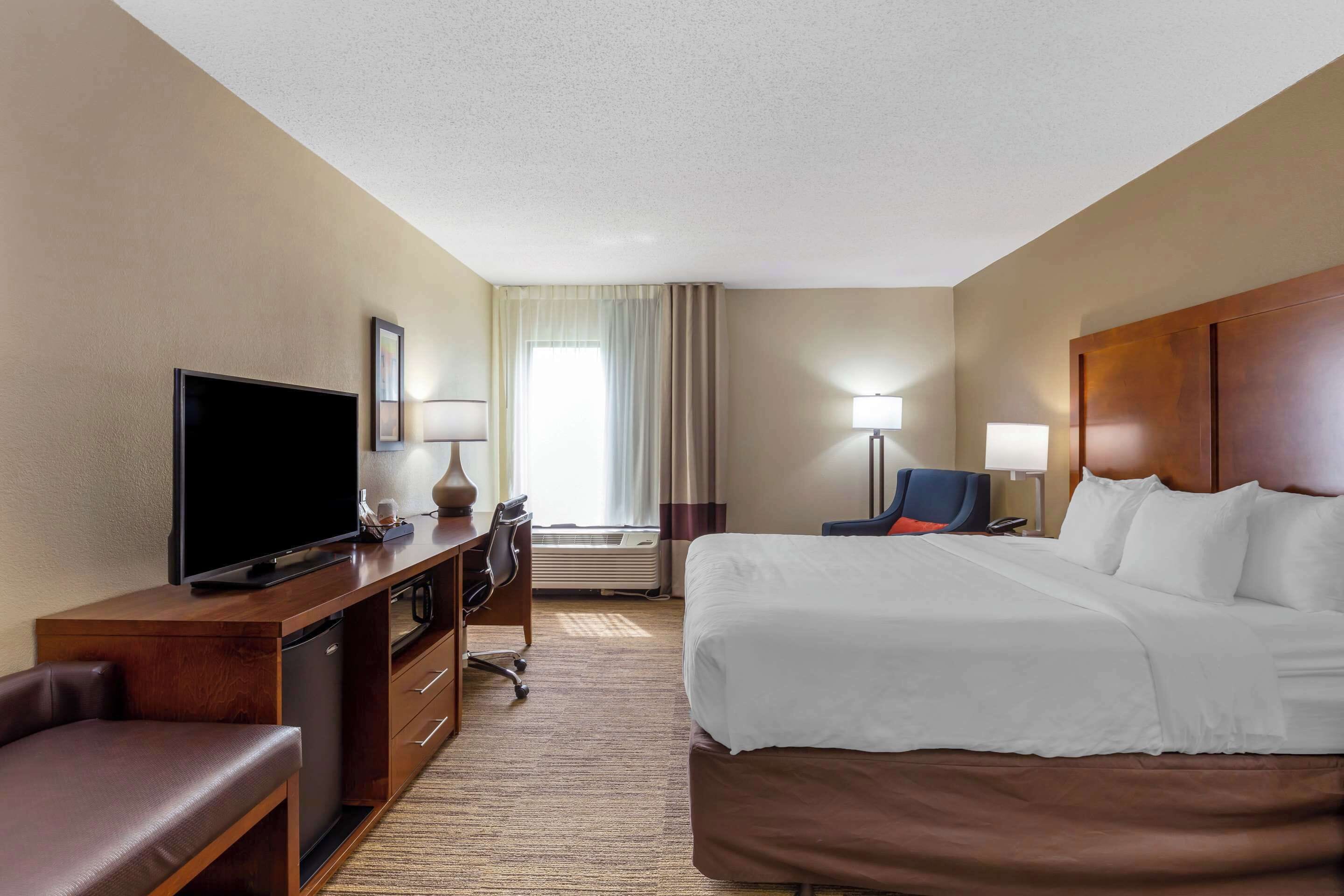 A hotel room with a bed, a desk with a television, a chair, and a desk lamp.